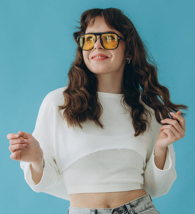 Jeune femme souriante aux cheveux bouclés, vêtue d’une tenue élégante, affichant une attitude confiante et joyeuse sur un fond bleu vif.