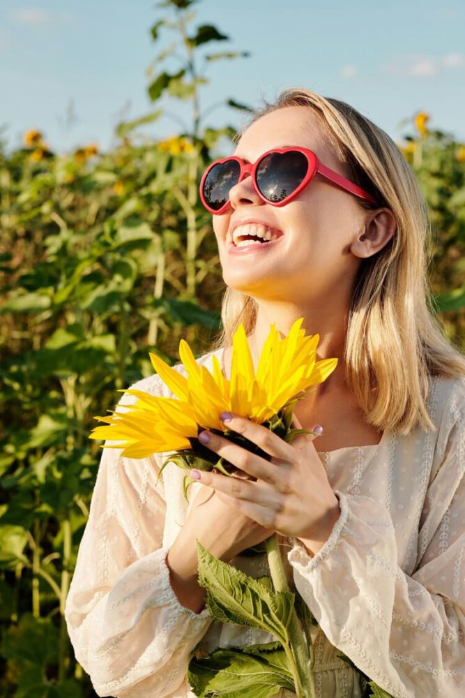 Une femme souriante portant des lunettes en forme de cœur tient un tournesol jaune vif dans un champ ensoleillé. Elle dégage du bonheur et de la chaleur sous un ciel bleu.