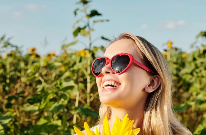 Une femme souriante portant des lunettes en forme de cœur tient un tournesol jaune vif dans un champ ensoleillé. Elle dégage du bonheur et de la chaleur sous un ciel bleu.