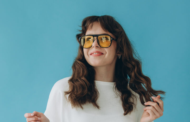Jeune femme souriante aux cheveux bouclés, vêtue d’une tenue élégante, affichant une attitude confiante et joyeuse sur un fond bleu vif.